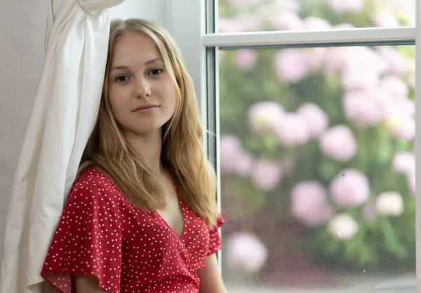 A young girl in a red dress sits on the floor in front of a bush with pink flowers and looks at the camera. High quality photo