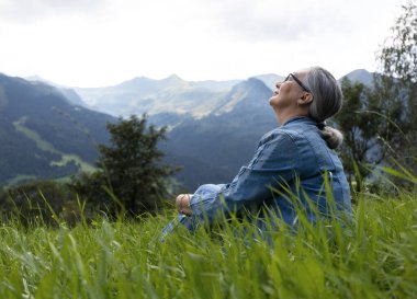 An elderly woman in a denim suit sits on a alpine meadow and looks at the sky. Gasteinertal, Salzburg, Austria. High quality photo