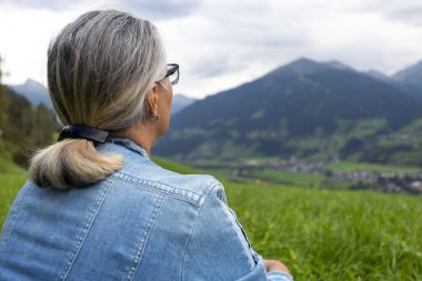 An elderly woman in a denim suit sits on a high meadow and looks at the mountains on the opposite side of the gorge. Austria, Salzburg. High quality photo