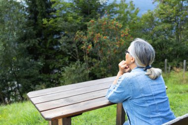A gray-haired woman sits at a wooden table in the countryside at the edge of a forest. Austria, Salzburg. High quality photo