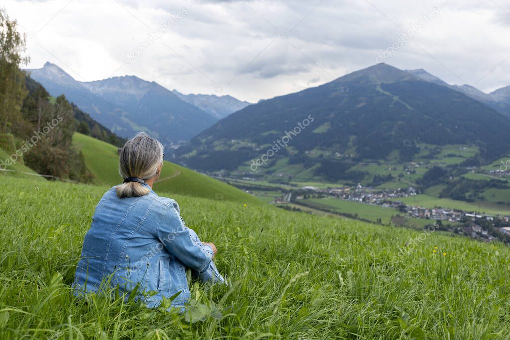 A gray-haired woman in a denim suit sits on a grassy slope and looks towards the mountains. Austria, Salzburg. High quality photo