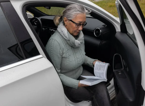 An adult gray-haired woman in a warm sweater sits in a car and reads the instructions. High quality photo
