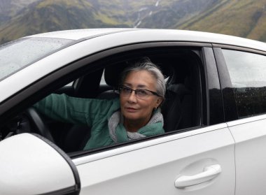 Gri saçlı bir bayan açık pencereli bir yolcu aracının direksiyonunda oturur ve kameraya bakar. Yüksek kalite fotoğraf
