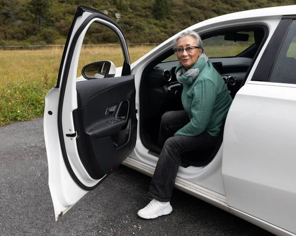 Gri saçlı bir kadın arabadan inip kameraya gülümsüyor. Yüksek kalite fotoğraf