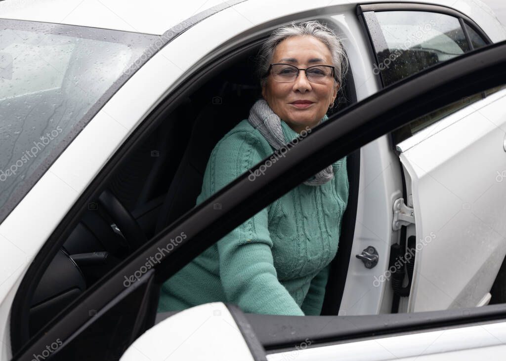 A gray-haired lady sits on the drivers seat in a car with an open door and smiles at the camera. High quality photo