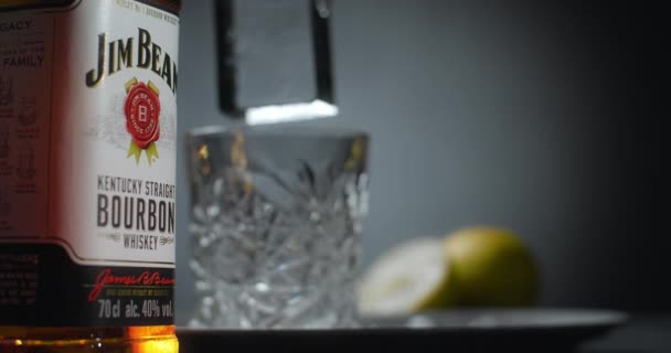 WARSAW, POLOGNE - 10 SEPTEMBRE 2021 : Un grand glaçon est déposé dans le verre de whisky au ralenti avec la bouteille de Jim Beam bourbon en arrière-plan, 4k Prores HQ 120 ips — Video