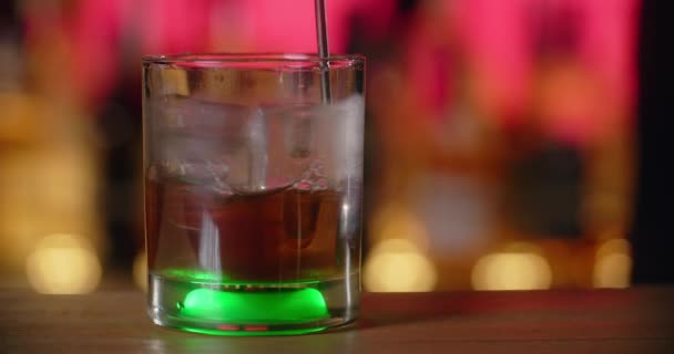 Bartender mengaduk minuman dingin dengan es batu dengan sendok berpegangan lama di bar, membuat koktail di meja bar, gelas koktail dengan lampu belakang, 4k Prores HQ 120 fps — Stok Video