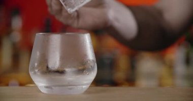 Barmen, dondurulmuş kokteyl bardağına forseps ile büyük buz küpü koyar, bu da kokteyli tezgahta yapar, 4k Prores HQ.