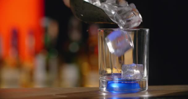 El camarero añade cubitos de hielo al vaso con retroiluminación azul en cámara lenta, lo que hace que el cóctel en el mostrador del bar, 4k Prores HQ 120 fps — Vídeo de stock
