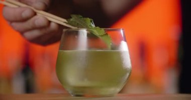 Barmen kokteyl bardağına taze şiso yaprağı ekliyor. Kokteyli Asyalı perilla otuyla yapıyor. Barmen tezgahta soğuk içecekleri karıştırıyor.