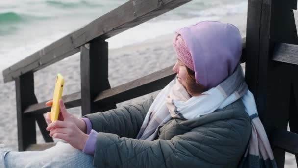 La blogger con ropa de abrigo hace selphie en la playa de invierno. Viajero en el mar frío en la temporada baja — Vídeo de stock