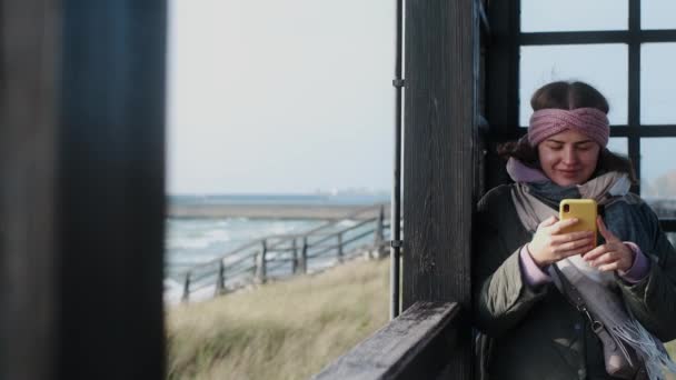 暖かい服で旅行ブロガーは、冬のビーチで彼女の携帯電話を使用しています, 4k 60p — ストック動画