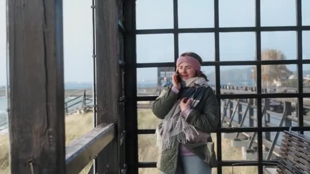 Mujer en ropa de abrigo sonríe y utiliza su teléfono en la playa de invierno, viajero en el mar en la temporada baja, 4k 60p — Vídeo de stock