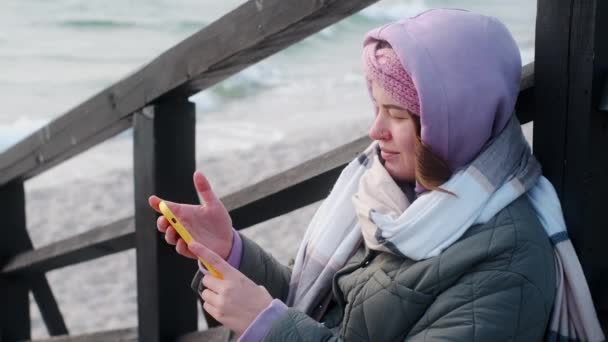 Τουριστική γυναίκα με ζεστά ρούχα χρησιμοποιεί το τηλέφωνό της στη χειμερινή παραλία, 4k 60p — Αρχείο Βίντεο
