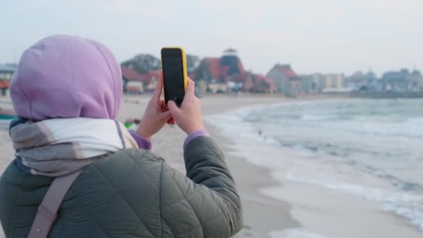 Blogerka w ciepłych ciuchach robi selfie na zimowej plaży. Podróżnik na zimnym morzu poza sezonem, 4k 60p — Wideo stockowe