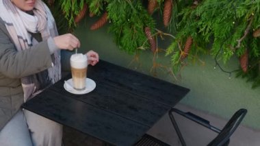 Güneş gözlüklü turist kadın, sonbaharda eski kasabanın sokak kafesinde sıcak kahve içer. Kadın latte 'yi uzun kaşıkla karıştırıyor, 4k 60 peni ağır çekimde.