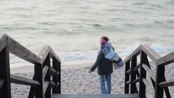 Ταξιδιώτισσα με ζεστά ρούχα κατεβαίνει στην παραλία κοντά στον παγωμένο ωκεανό. Τουριστική στην κρύα θάλασσα κατά την εκτός εποχής, 4k 60p Prores HQ — Αρχείο Βίντεο