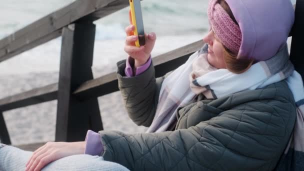 Γυναίκα blogger με ζεστά ρούχα κάνει selphie στη χειμερινή παραλία. Ταξιδιώτης στην κρύα θάλασσα εκτός εποχής, 4k 60p — Αρχείο Βίντεο