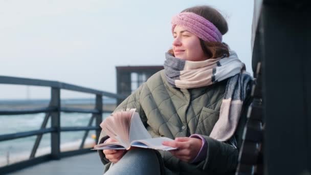 Γυναίκα με ζεστά ρούχα διαβάζει ένα βιβλίο στη θυελλώδη βόρεια θάλασσα. Traveller χαλαρώνει στη χειμερινή πλευρά της θάλασσας, 4k 60p — Αρχείο Βίντεο