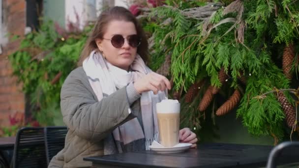 Turistka ve slunečních brýlích pije na podzim horkou kávu v kavárně starého města. Žena míchá latté po dlouhé lžíci, 4k 60p zpomalený pohyb — Stock video