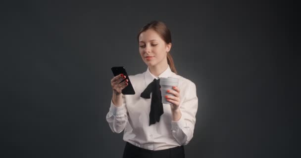 Ung kvinna dricker kaffe från återanvändbar kopp, med hjälp av smartphone och leenden, hon har arbetspaus på kontoret. 4k 120p Prores HQ — Stockvideo