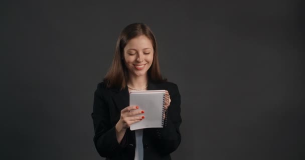 Young woman in black suit smiles and writes her ideas to the paper notepad, making the notes with happy look, 4k Prores HQ — Vídeos de Stock