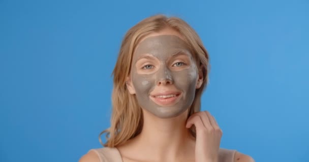 Blonde pretty woman poses in applied clay face mask. Womens cosmetics usage. Beauty care procedures on blue screen, 4k Prores HQ — Stock Video