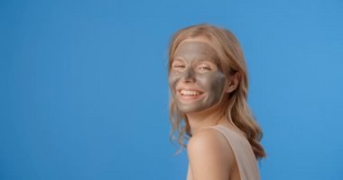 Smiling blonde woman poses in applied clay face mask. Womens cosmetics usage. Beauty care procedures on blue screen, 4k Prores HQ