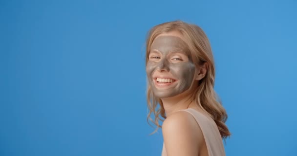 Smiling blonde woman poses in applied clay face mask. Womens cosmetics usage. Beauty care procedures on blue screen, 4k Prores HQ — Wideo stockowe