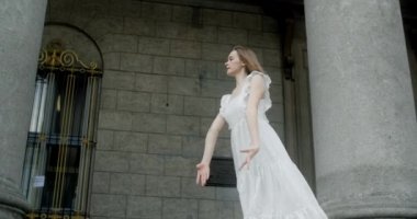 Young dancer in white dress dances on the stairs of the theatre in slow motion, balerina does dance steps and pa outdoors, 4k 120 fps Prores HQ