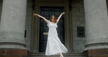 Young dancer in white dress dances on the stairs of the theatre in slow motion, balerina does dance steps and pa outdoors, 4k 120 fps Prores HQ