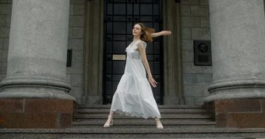 Young dancer in white dress dances on the stairs of the theatre in slow motion, balerina does dance steps and pa outdoors, 4k 120 fps Prores HQ