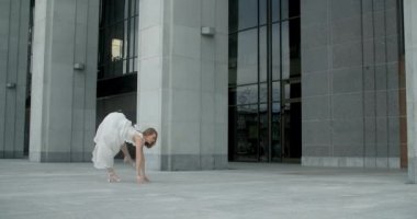 Young dancer in white dress does leg swing in slow motion outdoors, balerina dances in the gallery of columns architectural complex, 4k Prores HQ 120 fps