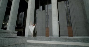 Young dancer in white dress does leg swing in slow motion outdoors, balerina dances in the gallery of columns architectural complex, 4k Prores HQ 120 fps