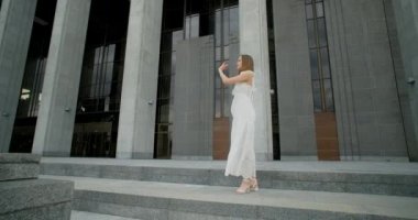 Young dancer in white dress does leg swing in slow motion outdoors, balerina dances in the gallery of columns architectural complex, 4k Prores HQ 120 fps