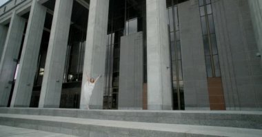 Young dancer in white dress does leg swing in slow motion outdoors, balerina dances in the gallery of columns architectural complex, 4k Prores HQ 120 fps