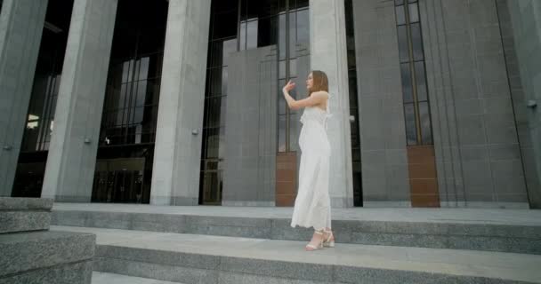 Young dancer in white dress does leg swing in slow motion outdoors, balerina dances in the gallery of columns architectural complex, 4k Prores HQ 120 fps — Vídeo de stock