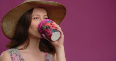 Woman in the summer dress and straw hat is drinking coffee and smiling on the pink backdrop, 4k Prores HQ 60p