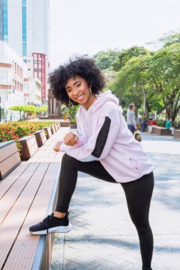 Afro saçlı Afro-Amerikalı genç bir kadın halka açık parkta spor yapıyor güneşli bir günde kameraya gülümsüyor.