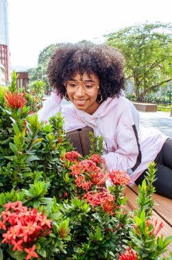 Güzel, genç, Afro-Amerikalı bir kadın. Bahçedeki çiçeklerle mutlu gülümsüyor. Halk parkında.