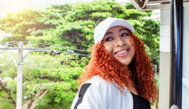 Binanın balkonunda gülümseyen kıvırcık saçlı güzel bir kız. Güneş ışığının altında ağaçların arkasında mutlu.