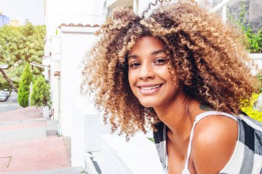 Güneşli bir günde, bahçede oturmuş kameraya gülümseyen kıvırcık kahverengi saçlı güzel bir afro kadın.
