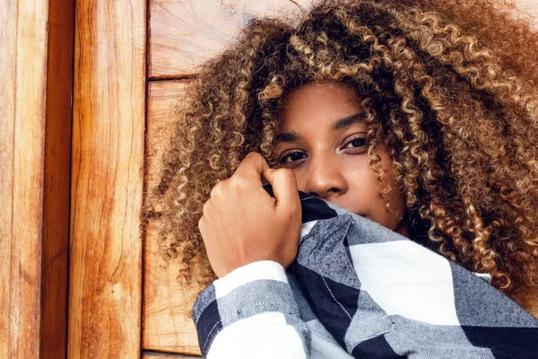 portrait of curly haired afro young teen woman in blue jacket covering her face in fashion on wooden background.