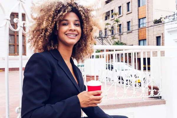 elegant business woman sitting in the city drinking coffee from her glass outdoors smiling, cheerful curly-haired afro woman drinking coffee.