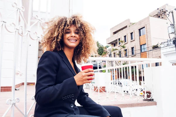 elegant business woman sitting in the city drinking coffee from her glass outdoors smiling, cheerful curly-haired afro woman drinking coffee.