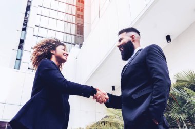 elegant curly-haired woman shaking hands with caucasian man with long beard making a deal outside business corporate building.