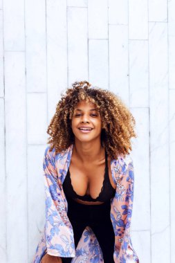 Güzel, kıvırcık saçlı, afro-amerikan kadın modaya uygun kıyafetleri ve kıyafetleri ile kentin beyaz tuğla duvarında mutlu bir şekilde gülümsemenin keyfini çıkarıyor..