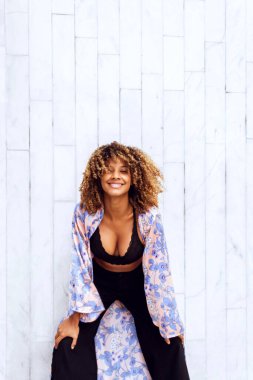 Güzel, kıvırcık saçlı, afro-amerikan kadın modaya uygun kıyafetleri ve kıyafetleri ile kentin beyaz tuğla duvarında mutlu bir şekilde gülümsemenin keyfini çıkarıyor..