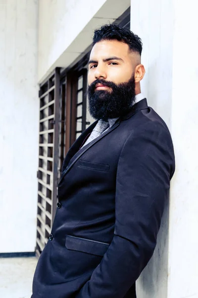 attractive successful man with long beard in elegant suit with tie leaning on wall looking at camera caucasian on white building walls