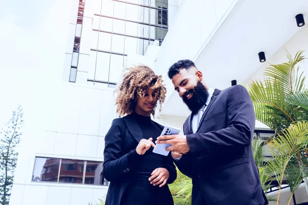 business couple outside corporate building. caucasian man with long beard showing his female partner something on his mobile having fun outdoors